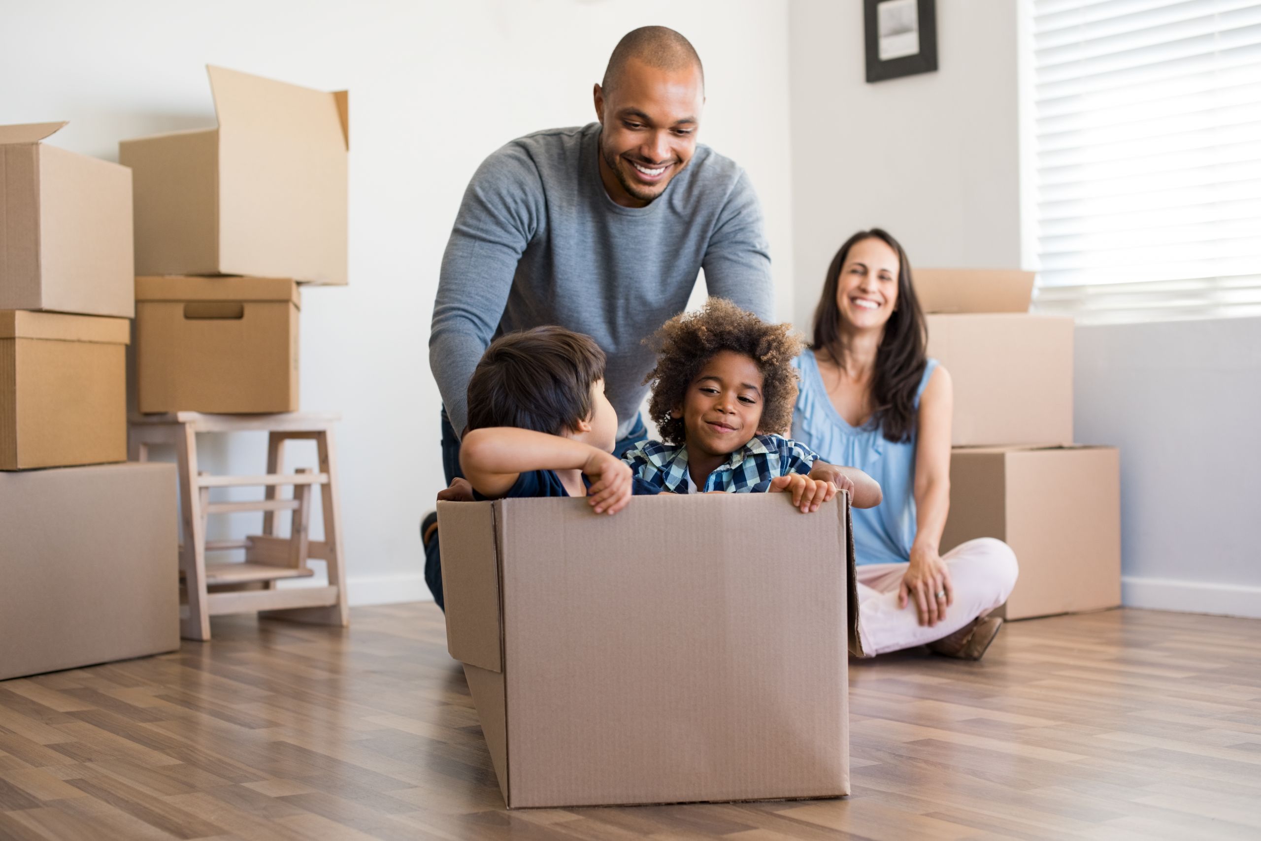 Happy african american father playing with children sitting in carton box at new home. Happy multiethinc family enjoying new home. Young parents and sons having fun during moving house.